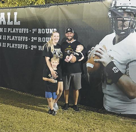 Luke Charles (right) with his wife Morgan (left) and his sons Jonathan Laith (left, in front) and Bennett Dean (right, in arms) in front of Luke’s Hall of Fame banner at UNC-Pembroke.