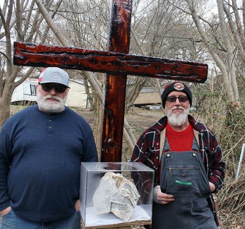 Dwight Otwell/Contributing Photographer Brothers Tracey (left) and Scotty Swann lost their homes in flooding from Hurricane Helene. Tracey was rescued by helicopter from his roof. Here, they stand beside the Bible that was lodged on a fence post. It is still on the fence post, but encased to safeguard it. Behind them is a wooden cross built by a South Carolina man, Joey Brown.
